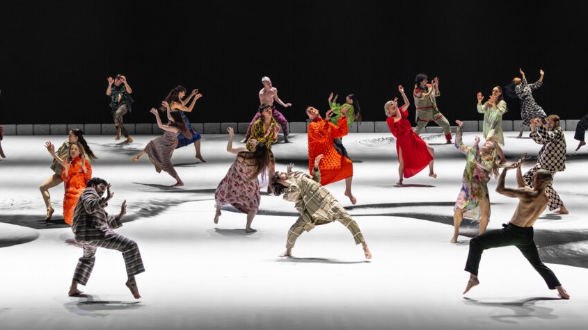 group of dancers in diverse, colorful costumes on a white and black stage, performing