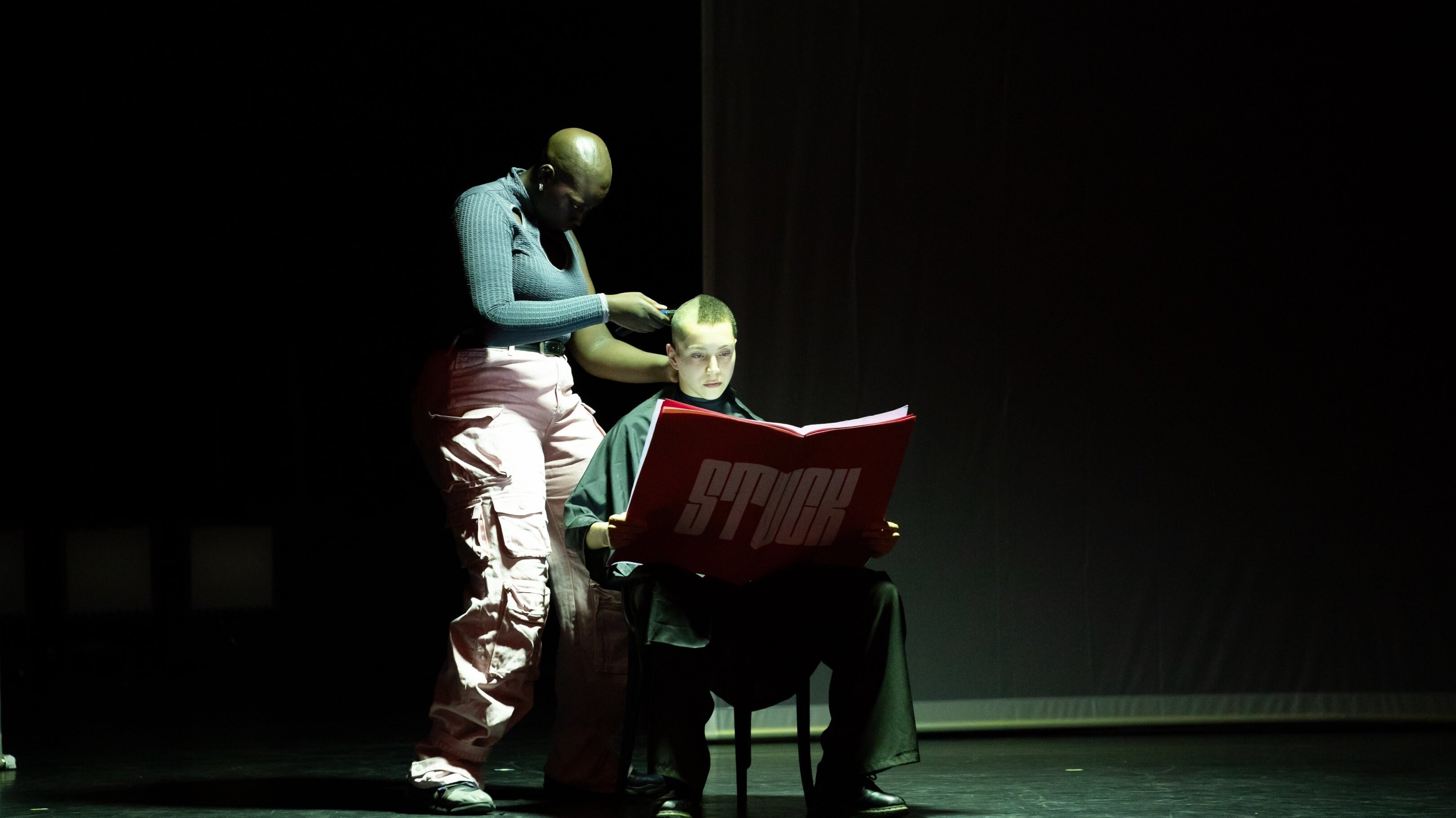one dancer shaves the head of another who is seated and reading a large red book with "STUCK" written on it
