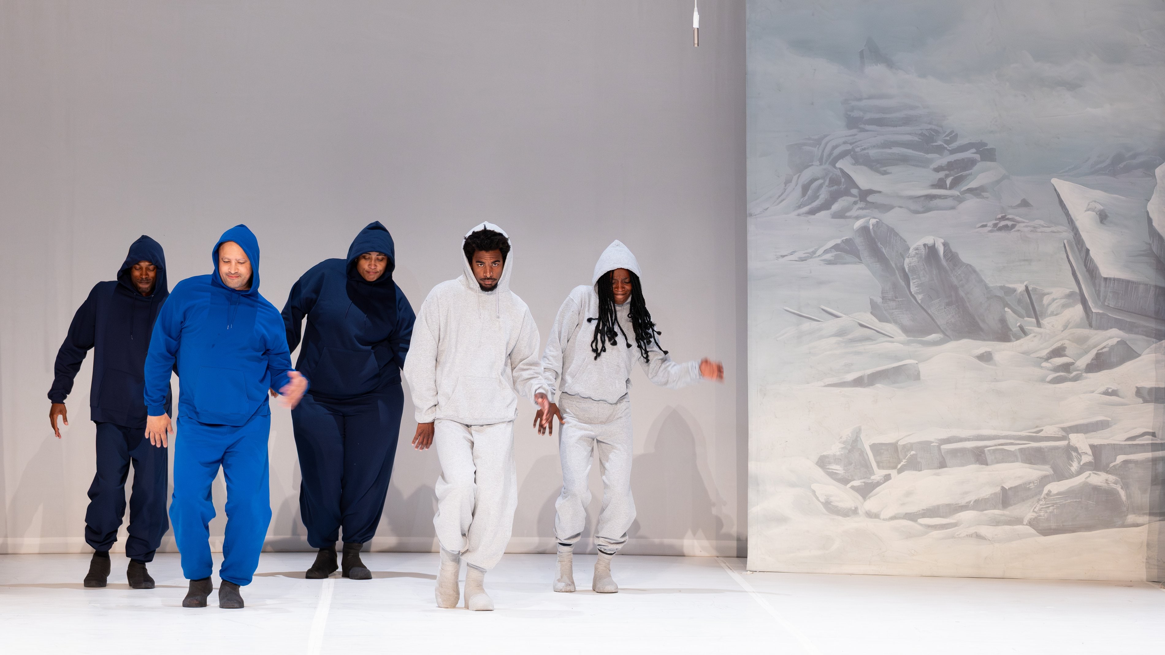 Cinq danseurs en survêtements bleu foncé, bleu vif, et gris clair, dansant en groupe, à droite de l'image un décor peint d'un paysage enneigé et rocheux