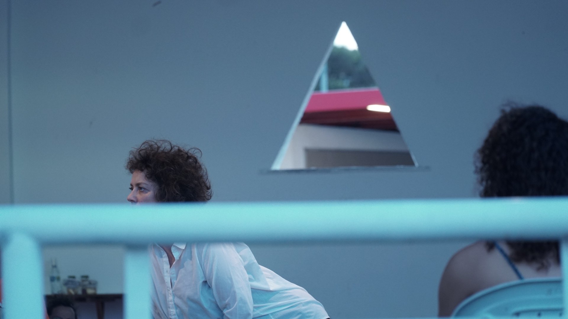Curly-haired woman in white shirt looking right, with triangular window and greenery in background