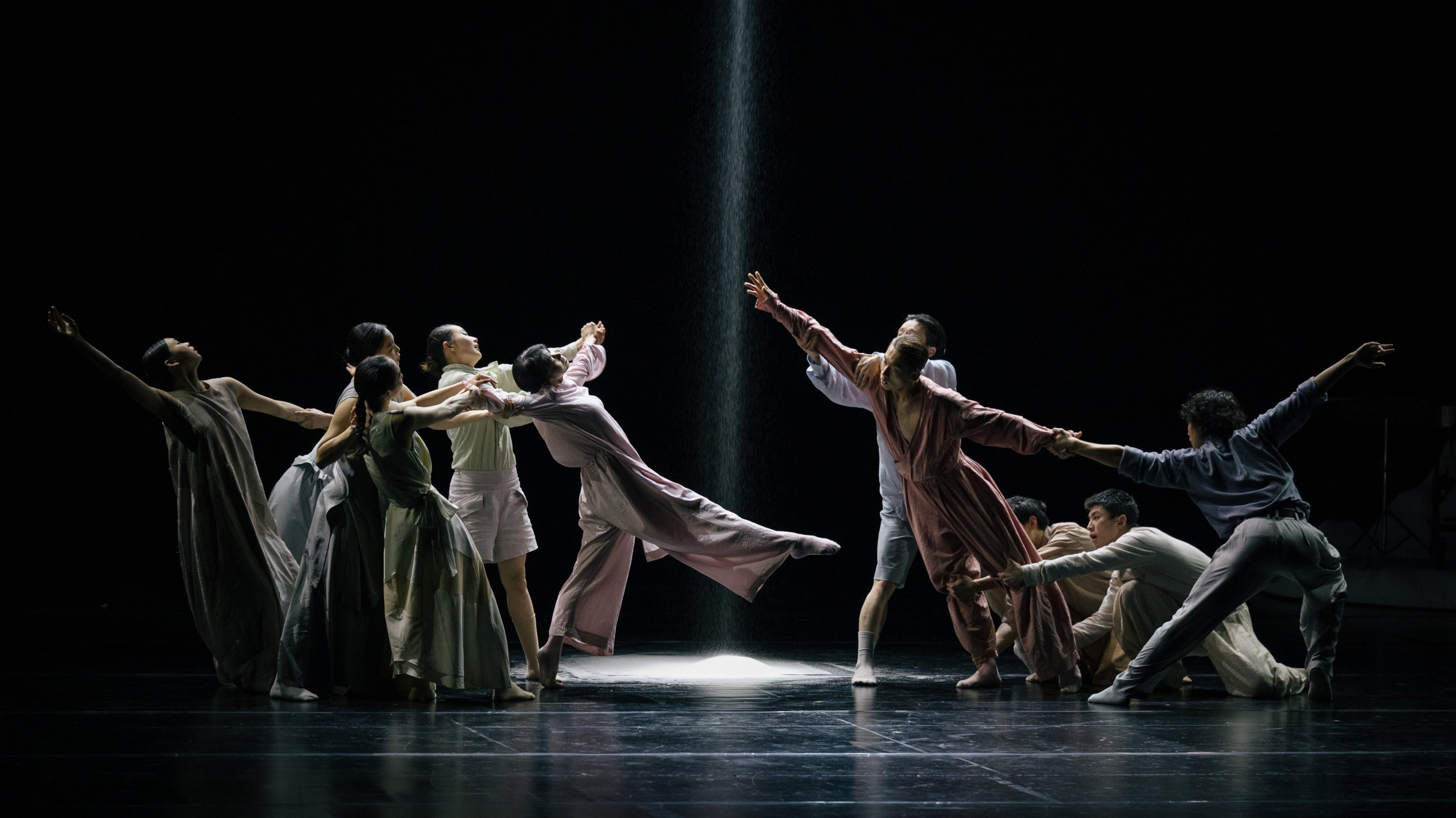 Dancers in flowing costumes on a dark stage, with a beam of light and falling sand in the center.