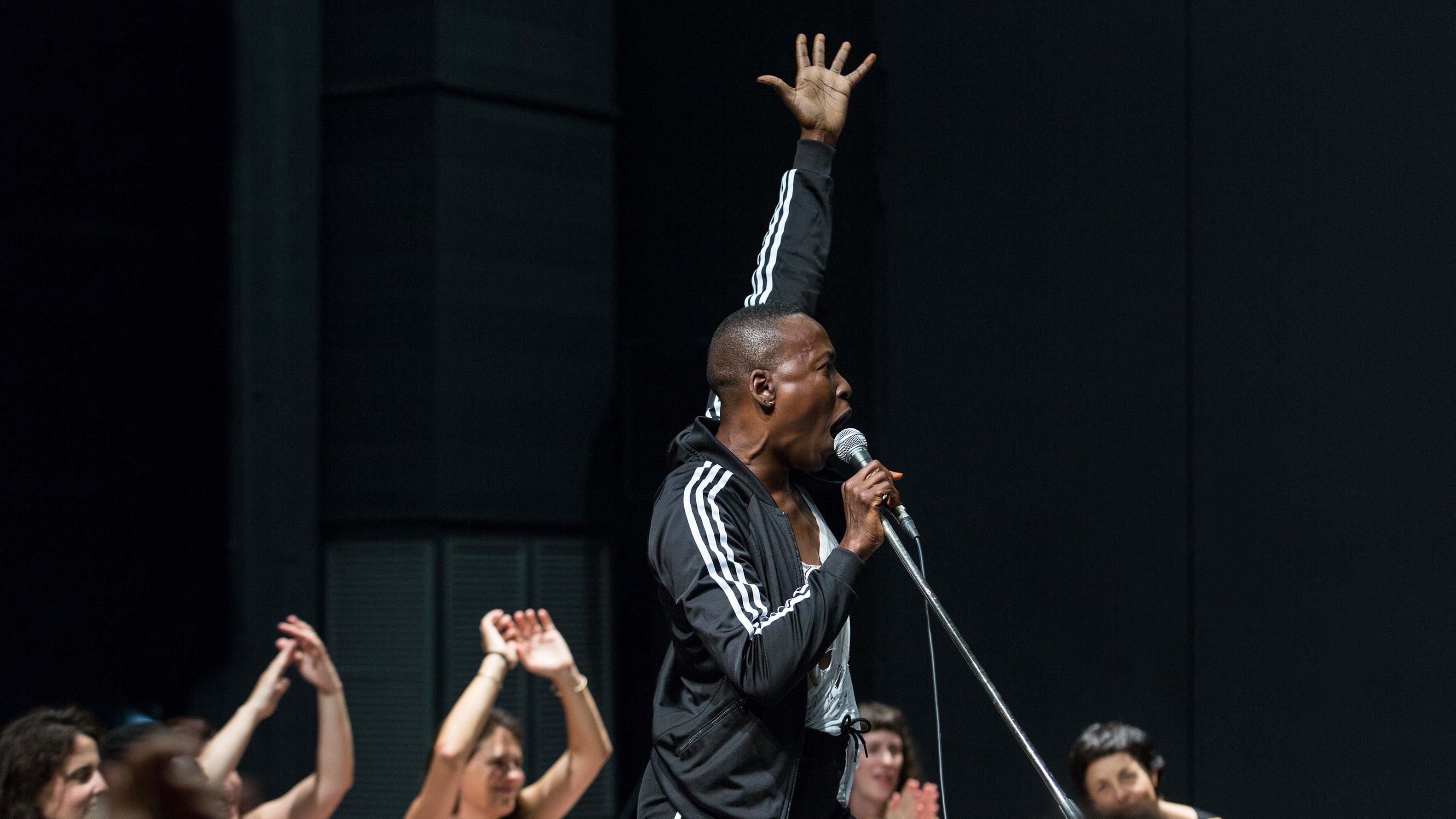 A performer in a black tracksuit sings passionately into a microphone, one arm raised, with an audience visible in the background, some with their hands raised