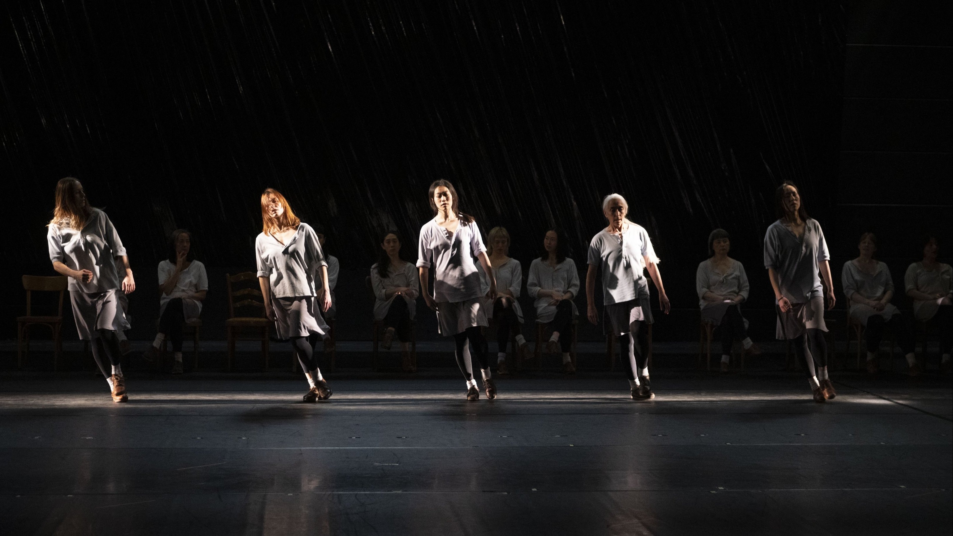 Dancers on a dark stage, some standing in the light, others seated in the shadow, wearing light tunics