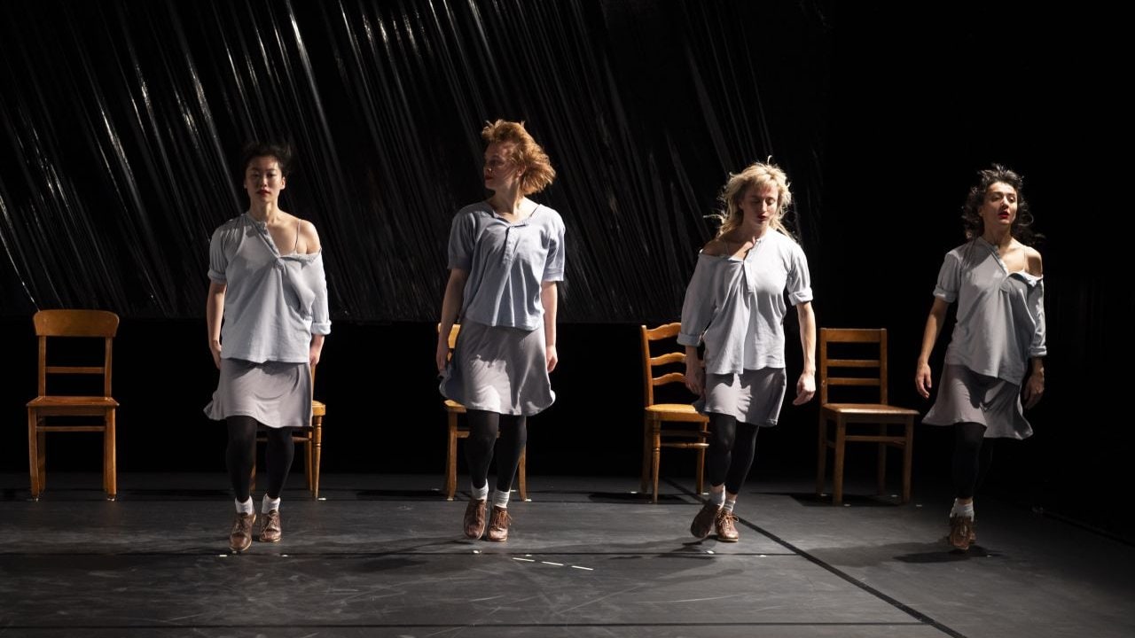 Quatre danseuses se tiennent debout alignées devant 4 chaises, avec un arrière fond noir. 