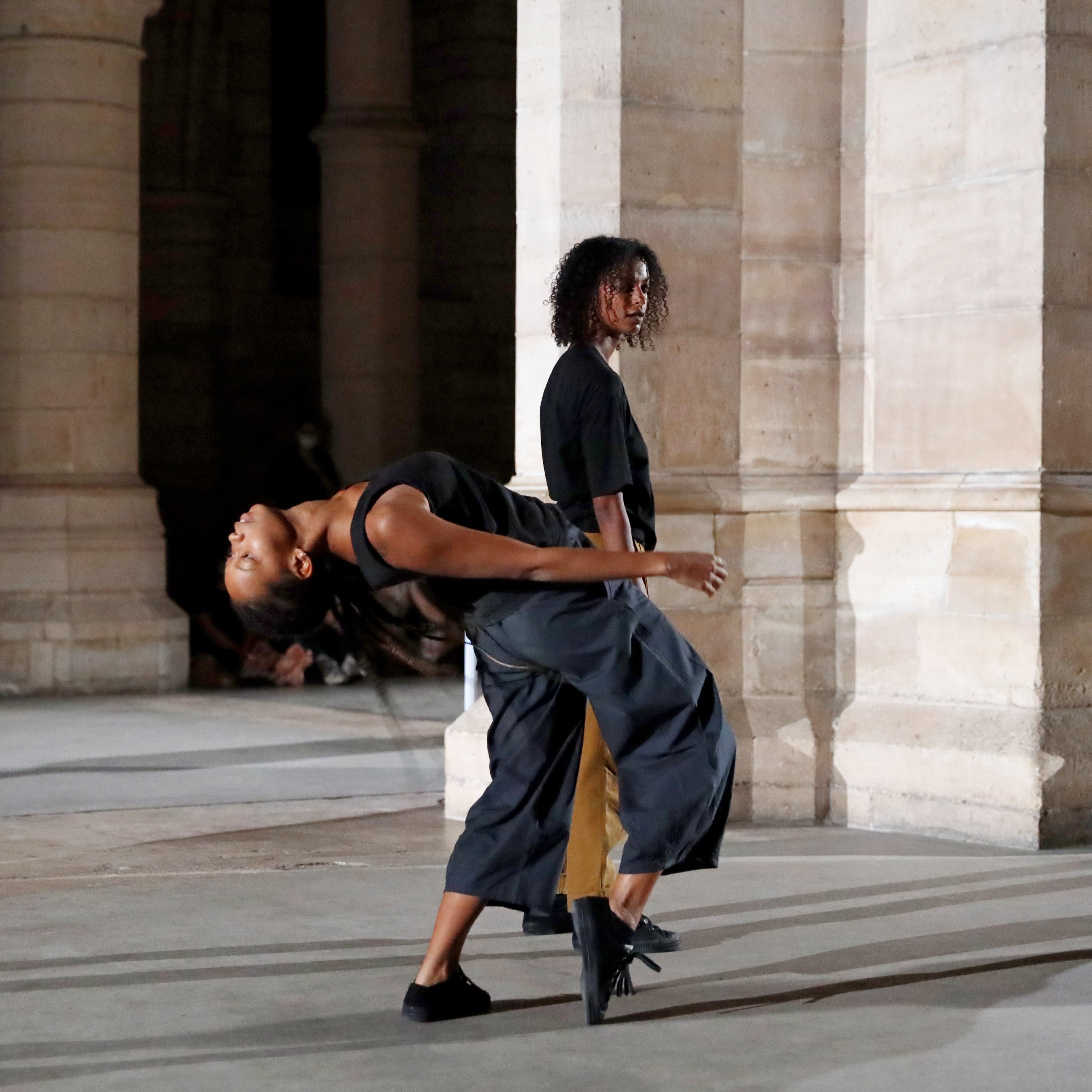 A Dancer standing behind another dancer bending backwards in a space with pillars