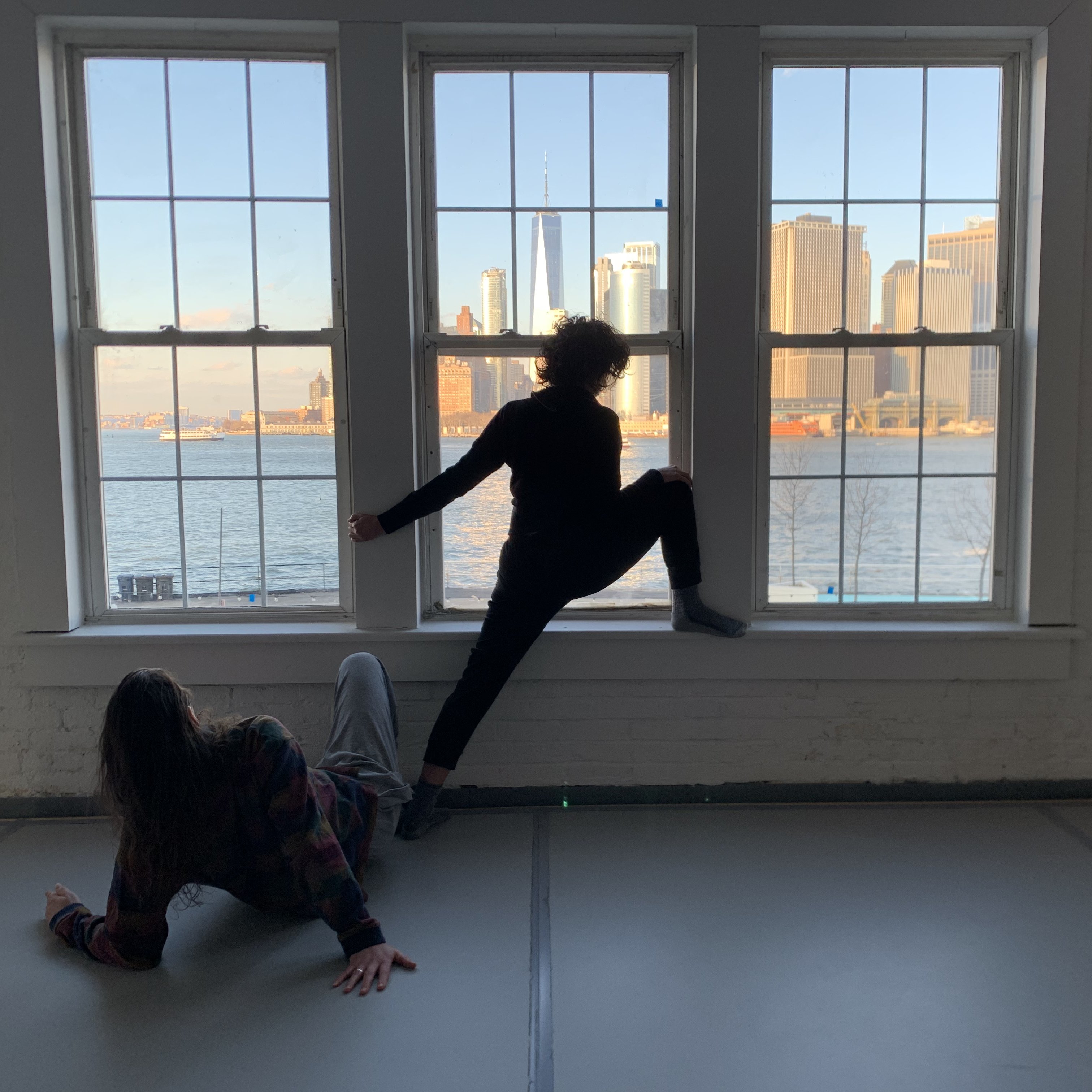 Two dancers stretching in front of a window with a view of the New York landscape