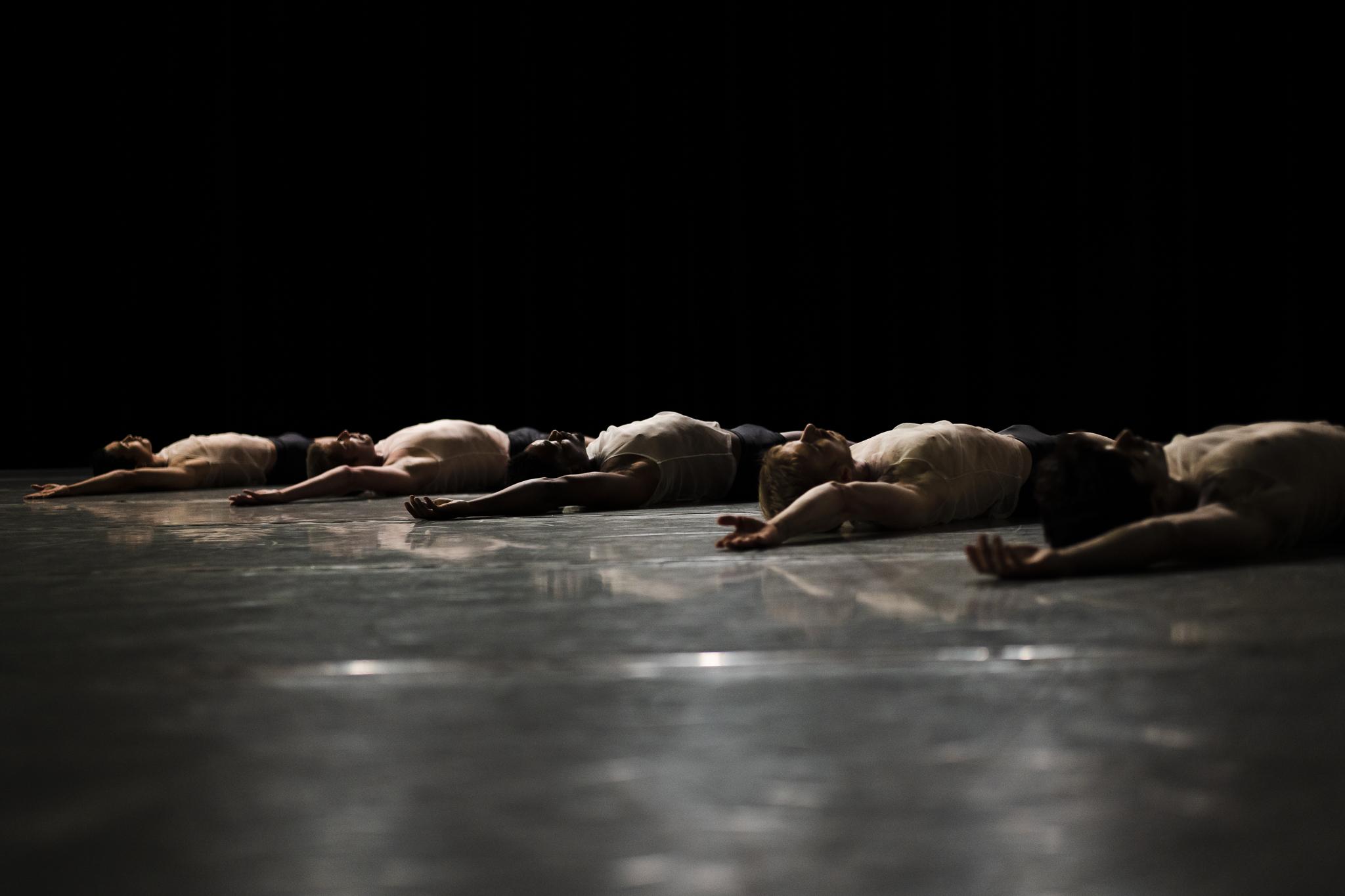 Dancers from the L.A. Dance Project lying on a black stage with their right arm raised.