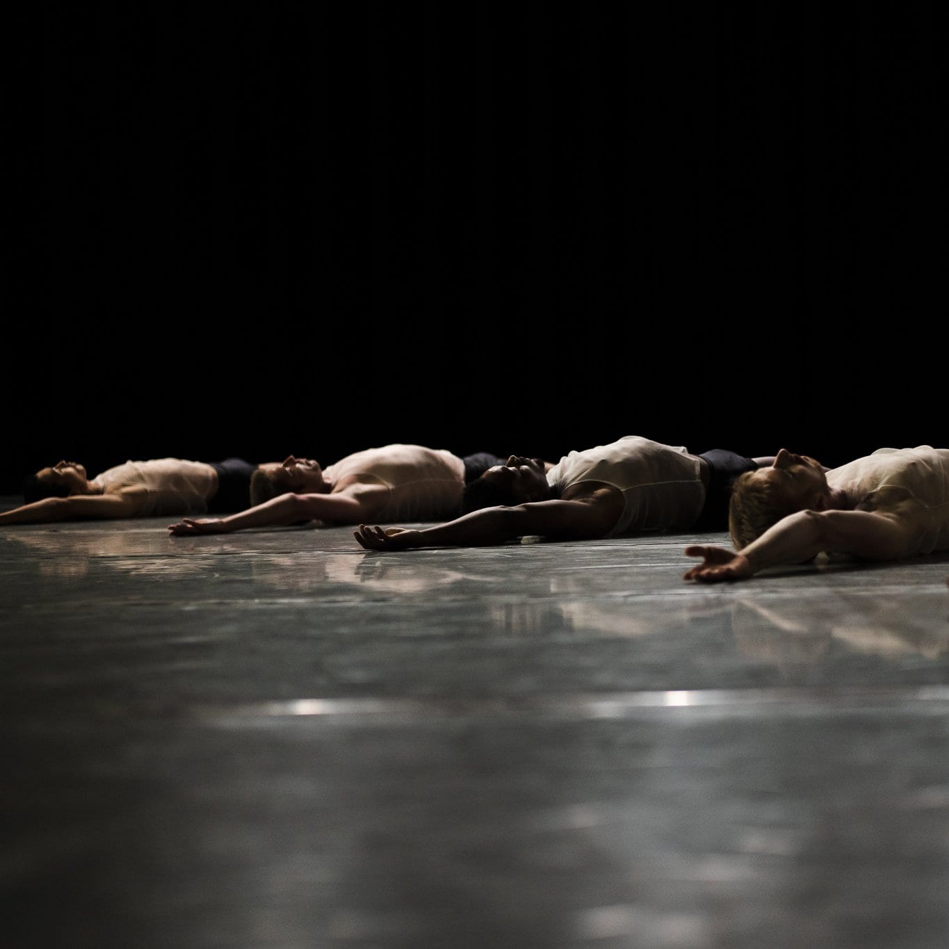 Dancers from the L.A. Dance Project lying on a black stage with their right arm raised.