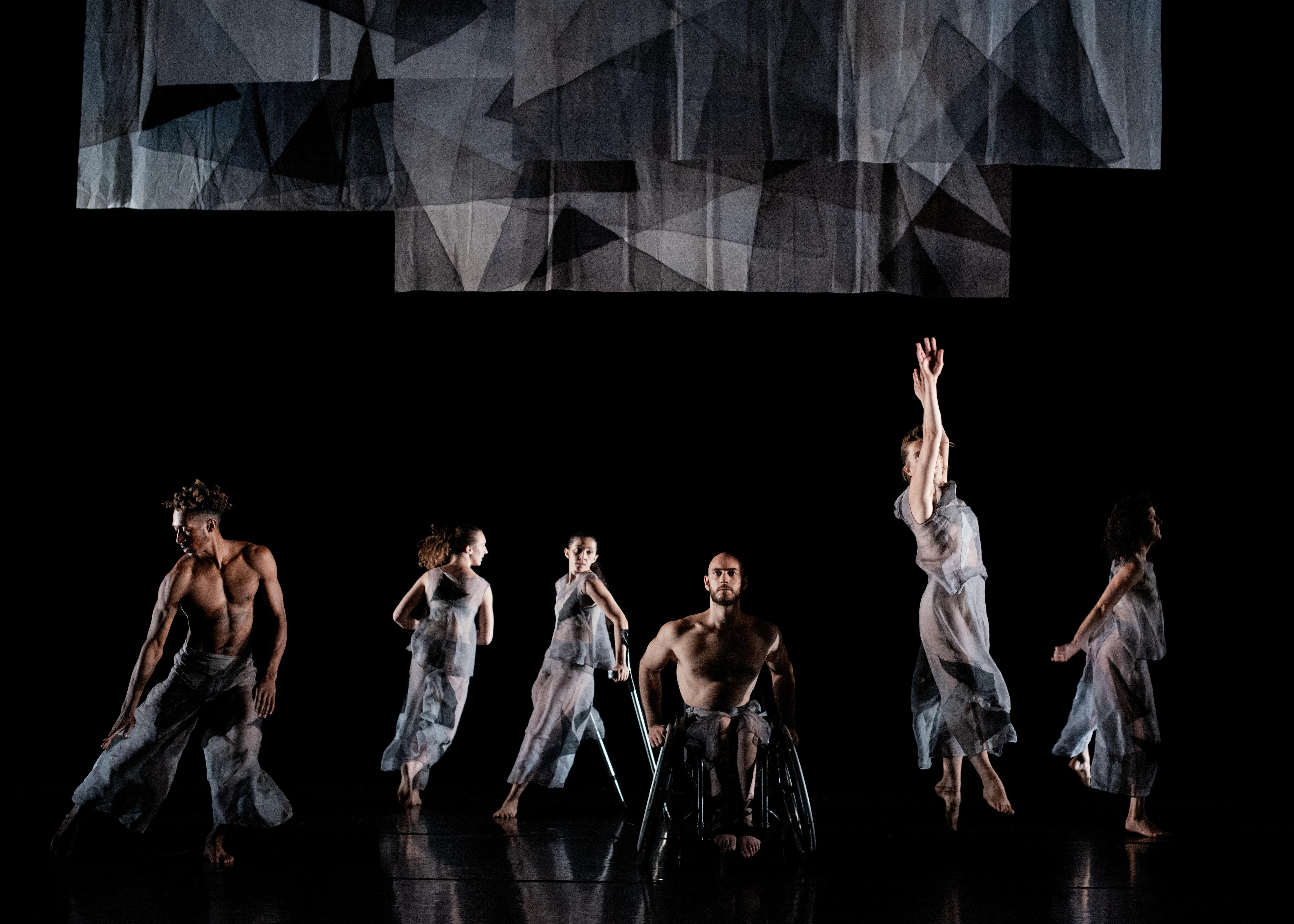 Six danseurs vêtus de vêtements transparents gris dansant sur une scène noire