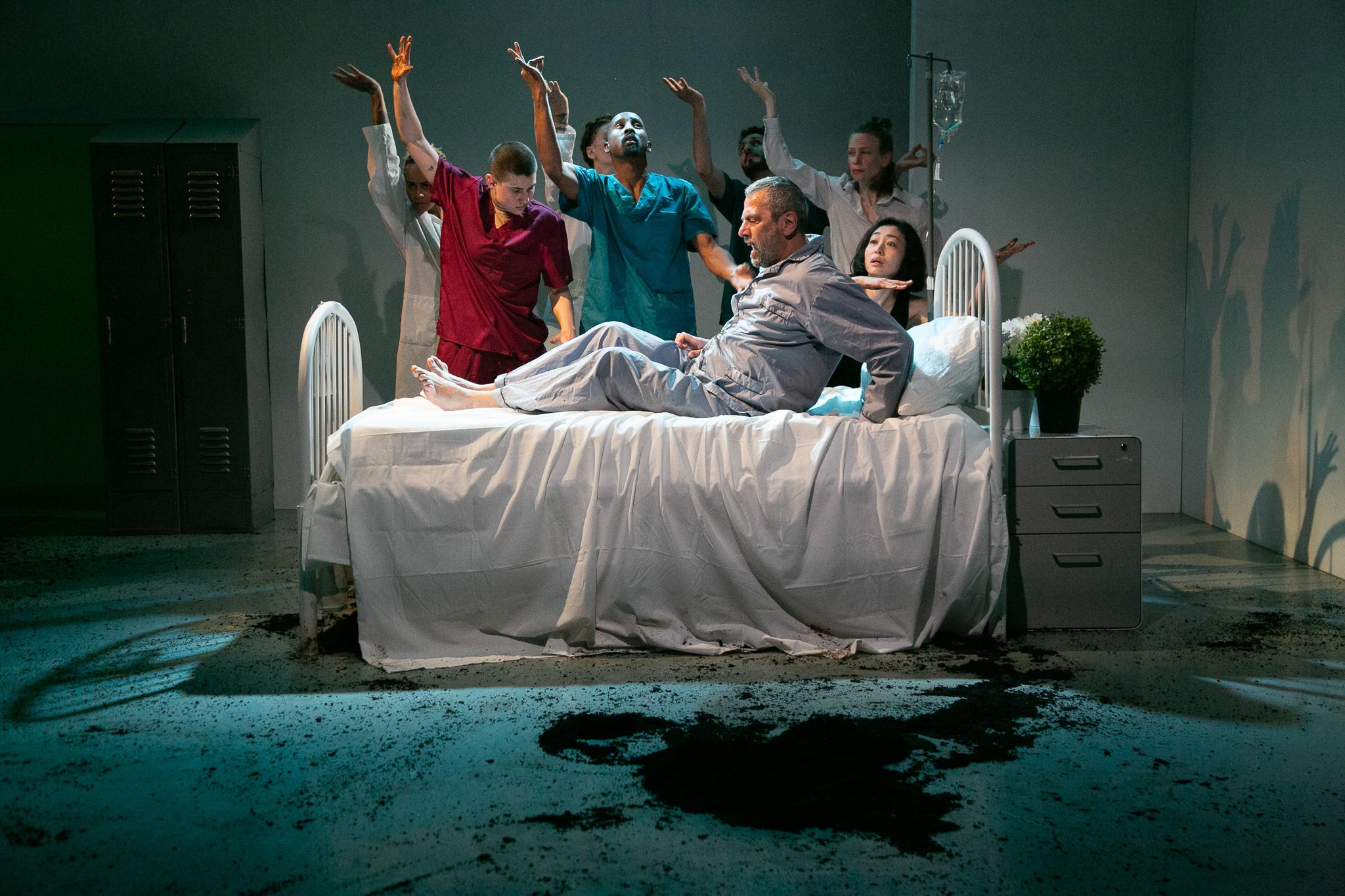 Dancers dressed as caregivers, surrounding a man on a hospital bed 
