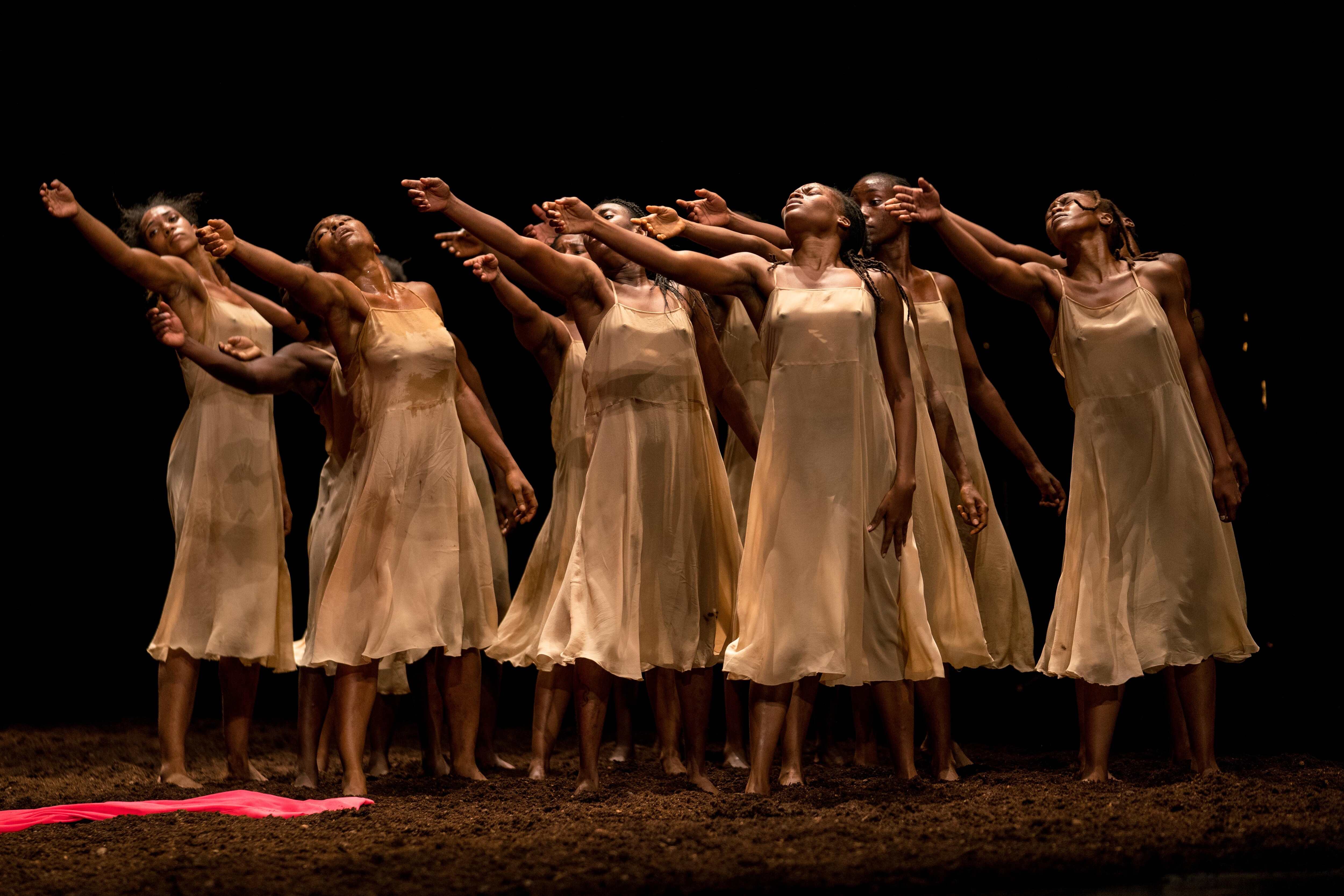 Women in white dresses, standing in dirt, facing three-quarters forward with their right arms raised.