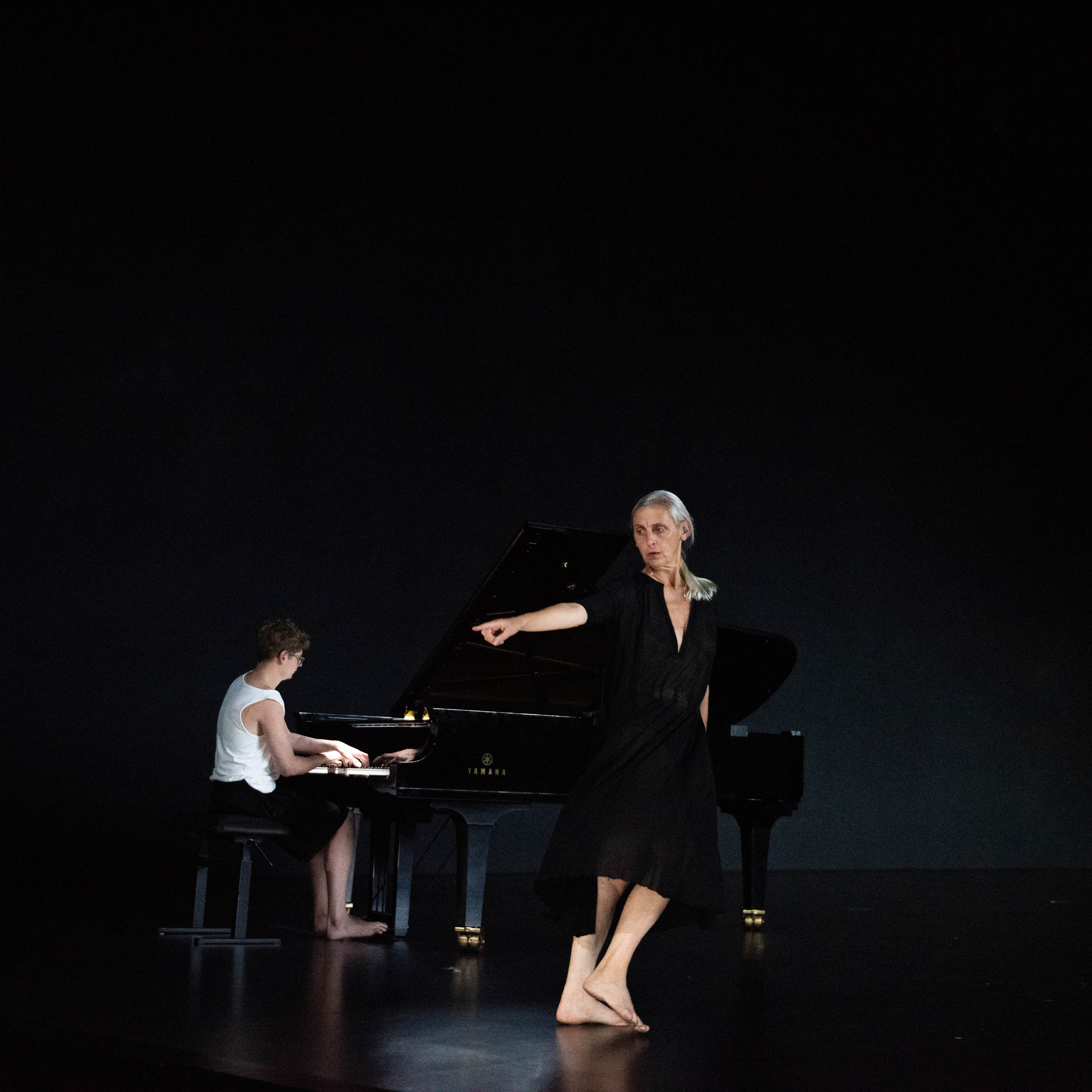 Sur une scène noire, une danseuse vêtue d’une robe noire avec un profond col en V pointe du doigt vers la gauche, ses pieds nus la menant dans la direction opposée avec, derrière elle, un jeune homme jouant du piano.