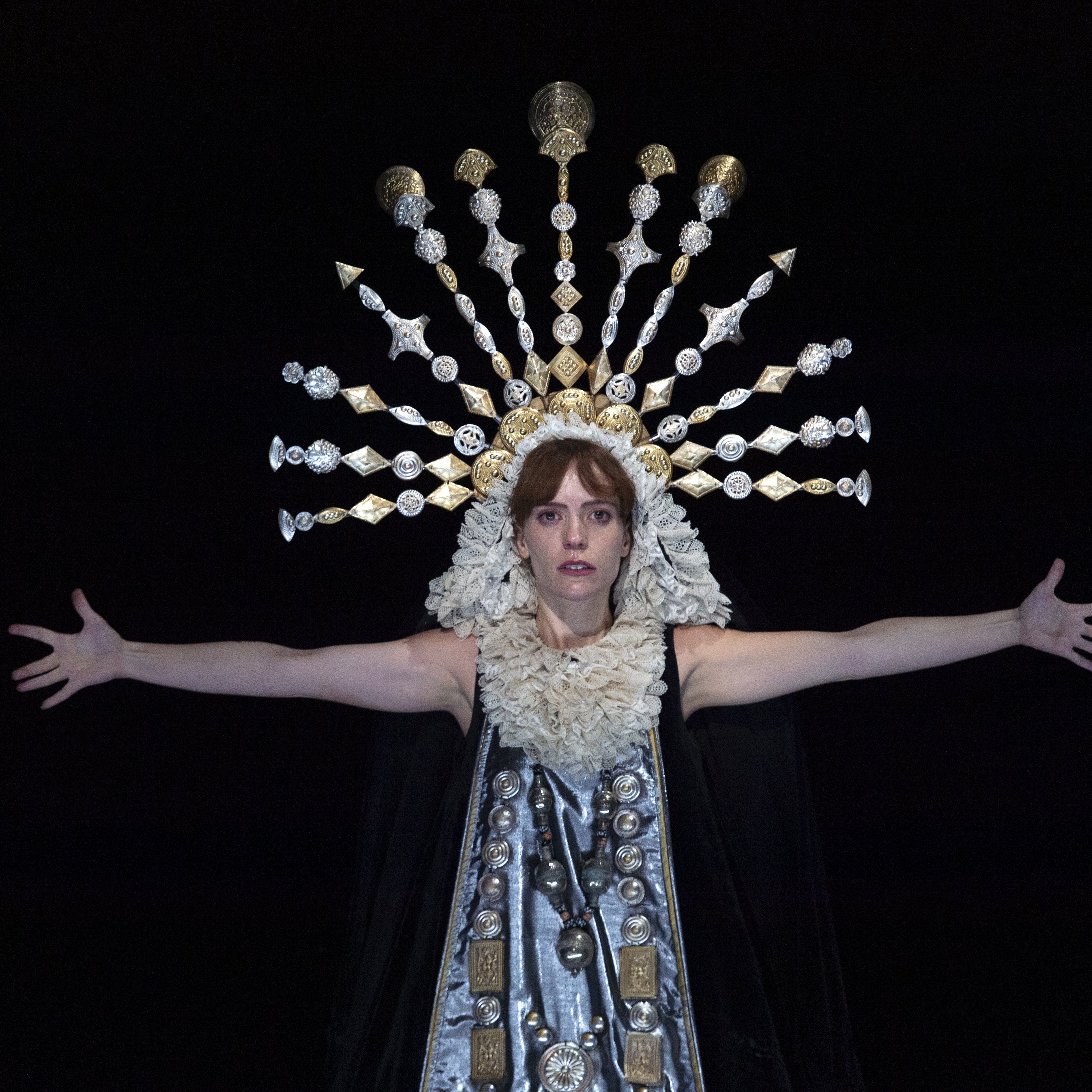 Woman in darkness whose face is bathed in light, wearing a crown in the shape of a halo, with her arms opened.