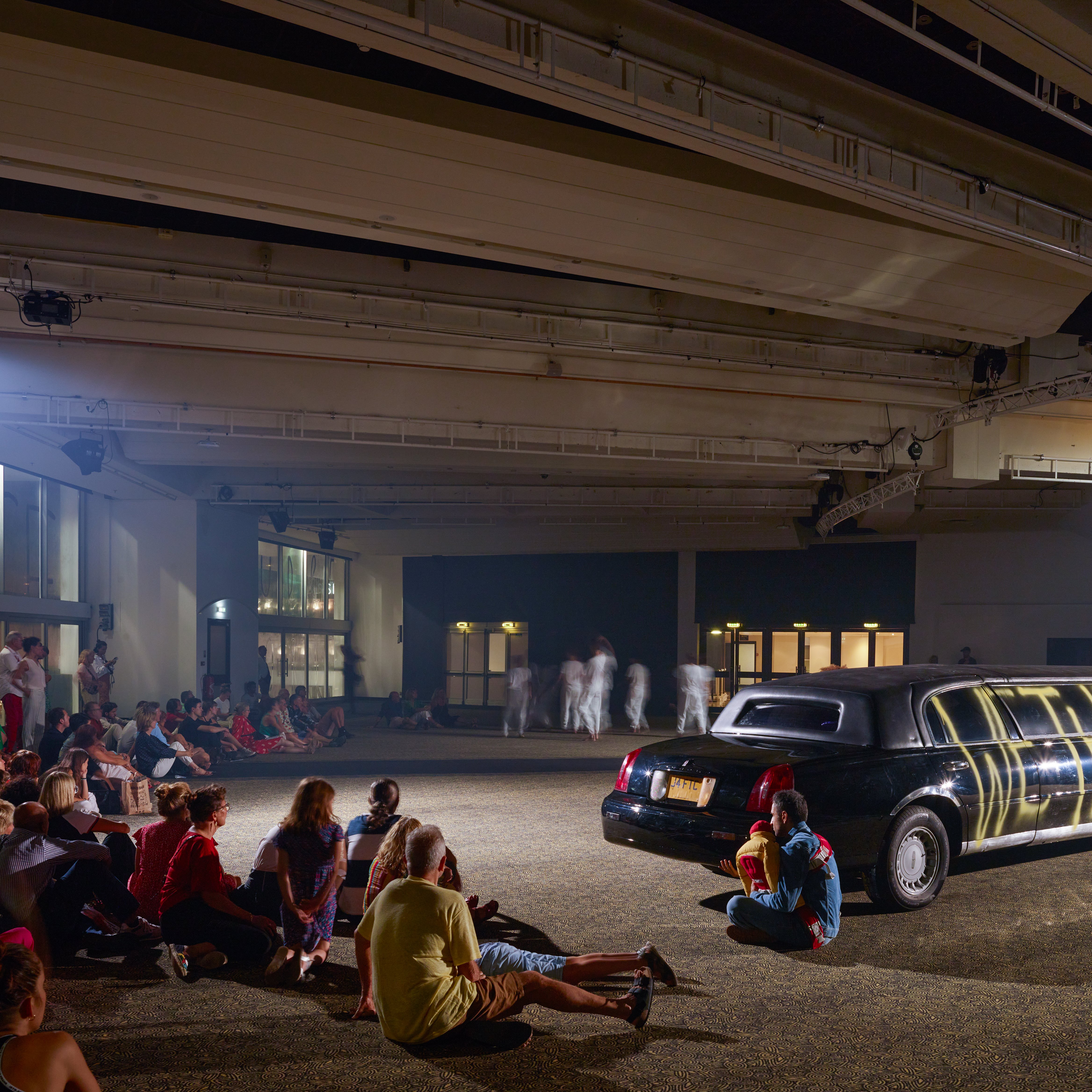 audience sitting on the floor in front of a black limousine