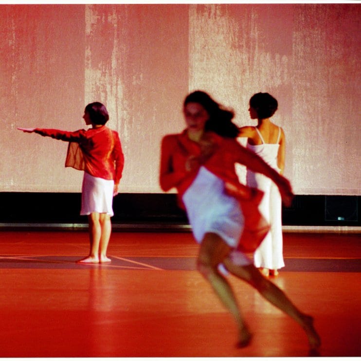 Trois danseuses sur scène, celle en premier plan court, les deux autres sont de dos en fond de scène, le bras gauche tendu à l’horizontal
