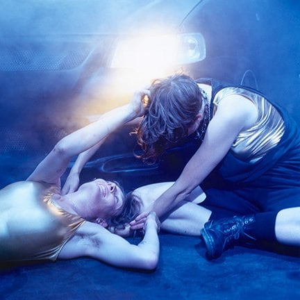 two dancers from Extra Life by Gisèle Vienne, one with his back to the floor, the other cross-legged, all in front of a car with its headlights on.